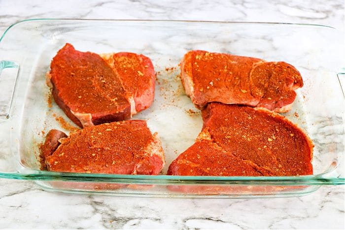 Four seasoned raw pork chops rest in a glass baking dish on a marble counter, waiting to rival the best grilled steak recipe you've ever tried.