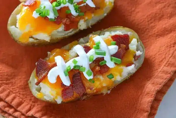 Two loaded baked potato halves rest on an orange cloth, each topped with sour cream, chopped bacon, melted cheddar cheese, and green onions. solid for make-ahead twice-baked potatoes, these savory delights are sure to impress at any meal.