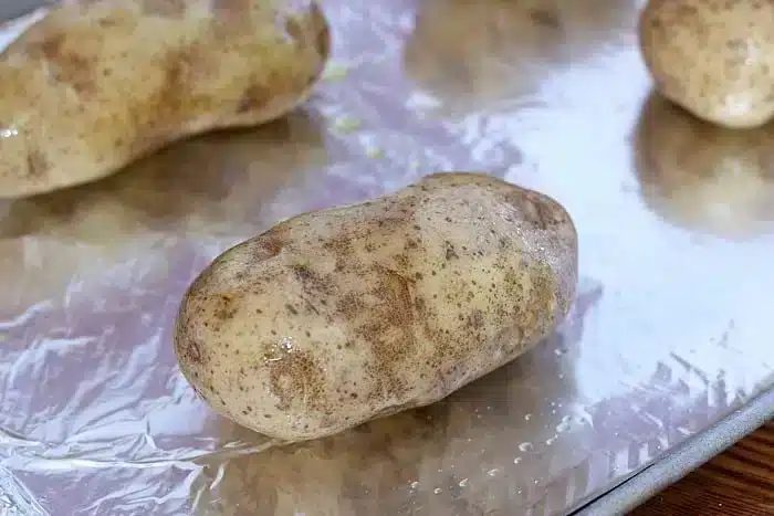 A large, raw potato coated in oil rests on a foil-lined baking sheet, solid for prepping Make Ahead Twice Baked Potatoes. Another potato peeks from the background, waiting its turn.