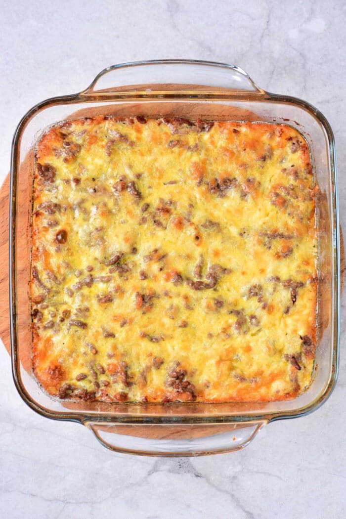 A Big Mac Casserole with a golden-brown cheese topping sits in a clear glass baking dish. The casserole, featuring ground meat and layers of melty cheese, boasts a slightly crispy surface. It rests invitingly on a white marble countertop, promising classic flavors in every bite.