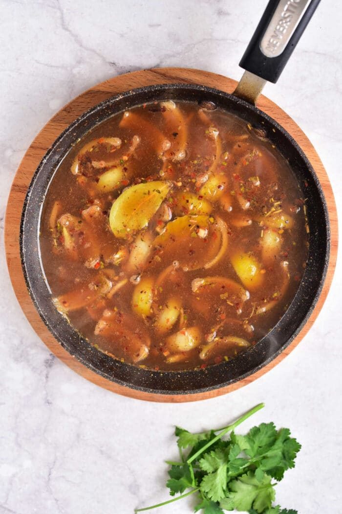 A skillet showcasing rich gravy and lemon slices sits invitingly on a wooden board, with fresh cilantro nearby on the marble surface, capturing the essence of a birria-inspired feast.
