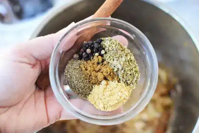 A hand holds a glass bowl of spices solid for birria tacos sauce, featuring whole black peppercorns, dried oregano, ground cumin, coriander, and ginger. In the background, a wooden spoon awaits to blend these flavors into a savory masterpiece.