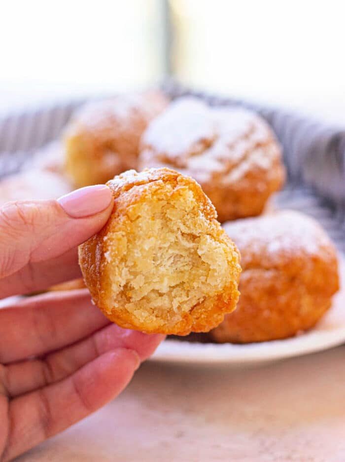 A hand delicately holds a partially eaten fried biscuit donut hole, with a plate piled high with similar treats tempting in the background.