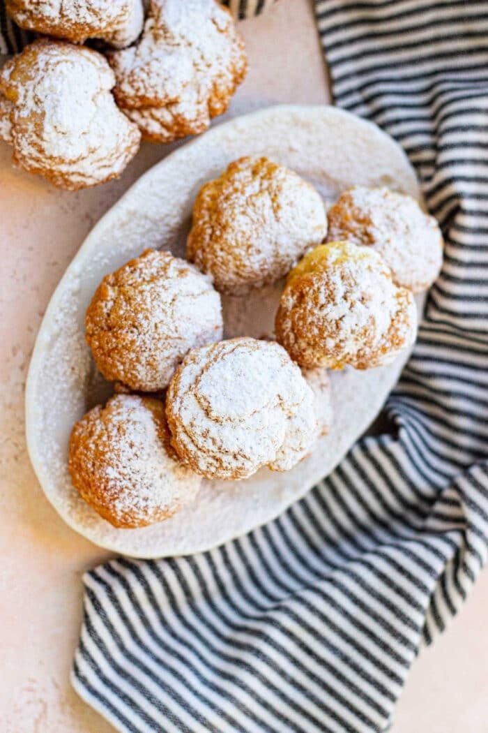 A white oval plate holds several powdered sugar-covered cream puffs, resembling delicate biscuit donut holes. More cream puffs are partially visible in the top left corner. The plate is placed on a light pink surface next to a black and white striped cloth.