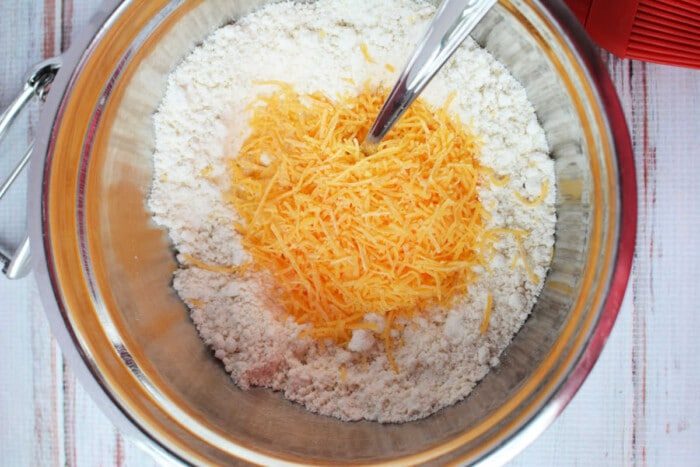 A mixing bowl filled with flour, shredded cheddar cheese, and a spoon rests on a wooden surface, resembling the solid start for savory biscuits using Jiffy Mix.
