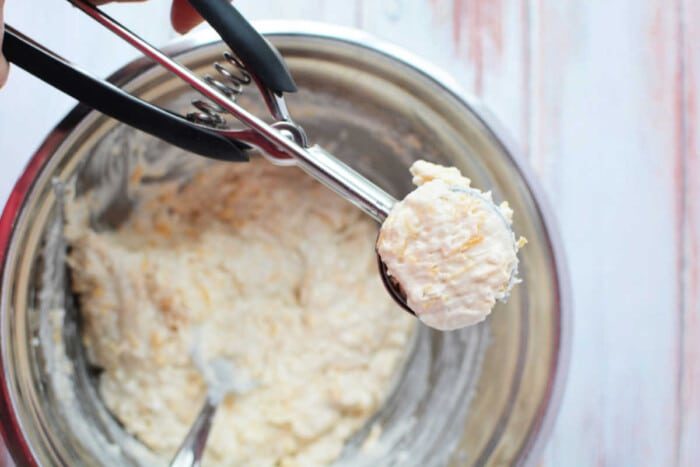 A cookie dough scoop filled with Jiffy Mix dough is held over a mixing bowl on a wooden surface, ready to bake good biscuits.