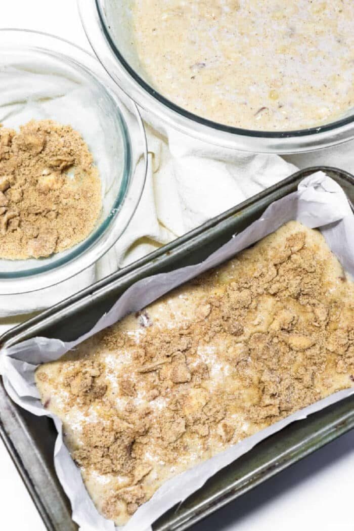 A loaf pan with Bisquick banana bread recipe batter topped with brown sugar, next to bowls of batter and brown sugar on a white cloth.