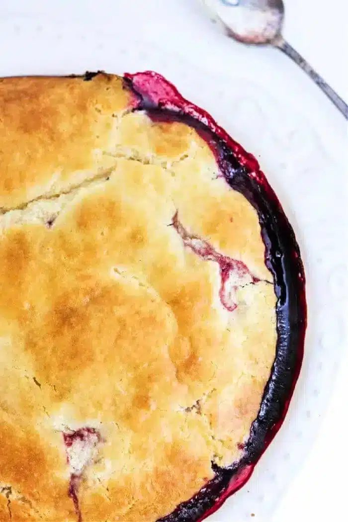 A close-up of a freshly baked bisquick pie with a golden-brown crust, slightly cracked, revealing a glimpse of dark red filling beneath, reminiscent of a blackberry cobbler. A silver spoon rests on a white plate beside the pie.