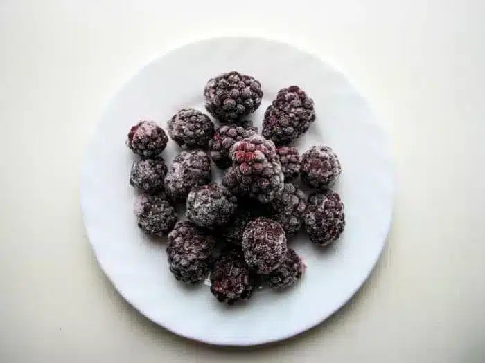 A white plate containing a small pile of frozen blackberries, solid for crafting a delightful blackberry cobbler, is centered on a light background.