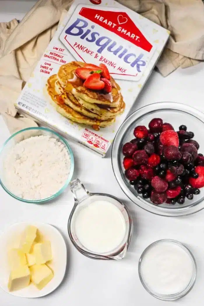 A box of Bisquick pancake mix is surrounded by bowls containing flour, mixed berries solid for a blackberry cobbler, a cup of milk, a dish of butter, and a container of sugar. The ingredients are arranged on a white surface with a beige cloth in the background.