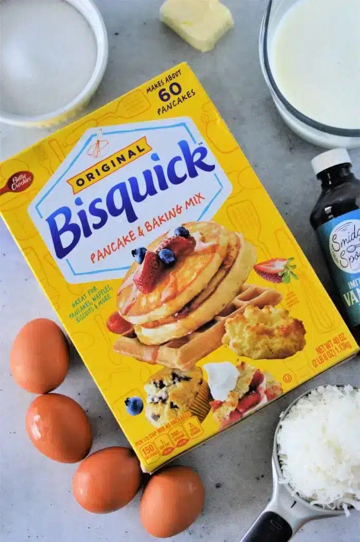 A box of Bisquick pancake and baking mix sits ready to create the famed Bisquick Impossible Pie Recipe, surrounded by three eggs, a bowl of milk, a block of butter, a bottle of vanilla extract, and a cup of shredded coconut on a gray surface.