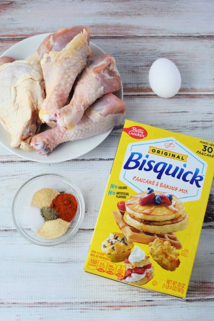 Ingredients for a mouthwatering oven-fried chicken recipe: raw chicken drumsticks, an egg, Bisquick mix, and spices mixed in a bowl on a rustic wooden surface.