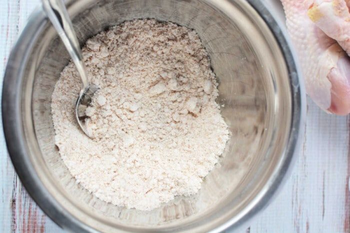 A bowl of Bisquick flour with a spoon nestled inside sits beside raw chicken pieces, ready to be transformed into crispy oven-fried chicken on a light wooden surface.