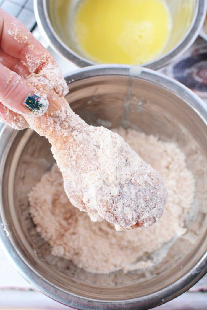 A hand expertly coats a chicken drumstick in Bisquick over a mixing bowl, preparing it for good oven-fried chicken.