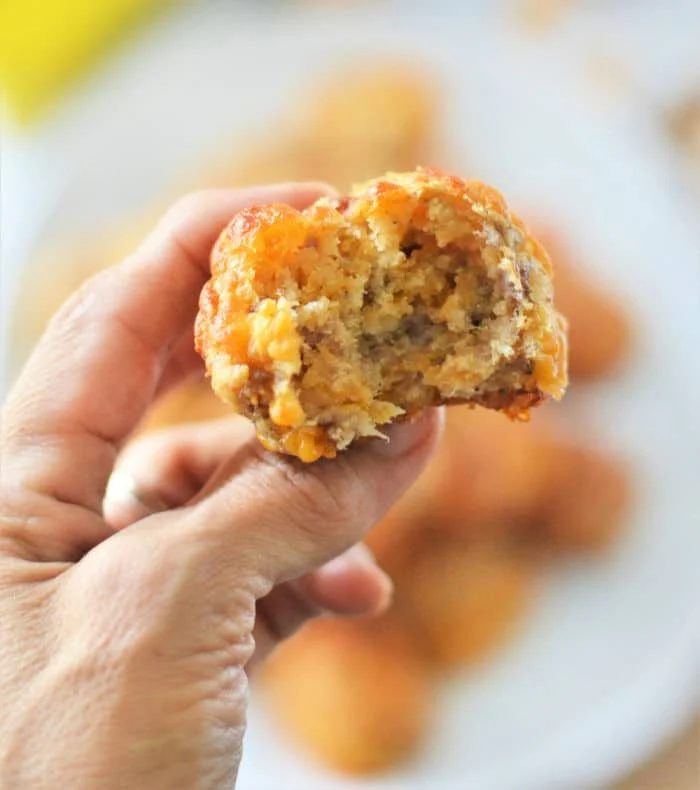 A hand holds a bitten savory Bisquick sausage balls, with a blurred background showcasing similar muffins on a plate.