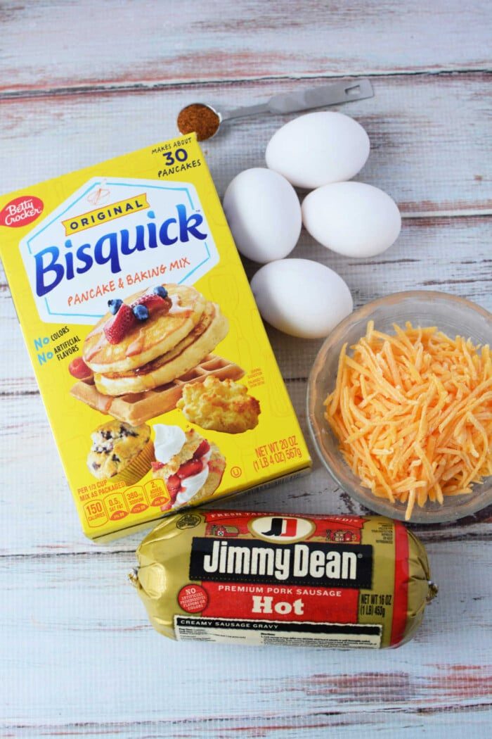 A Bisquick box, three eggs, shredded cheese, a hot sausage package, and a teaspoon of spice sit ready on a white wooden surface, solid for whipping up savory muffins.