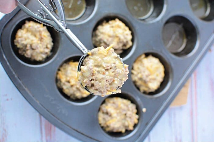 A scoop of muffin batter overflows a muffin pan filled with good muffins, ready to bake on a light wooden surface, much like preparing a savory Bisquick sausage treat.