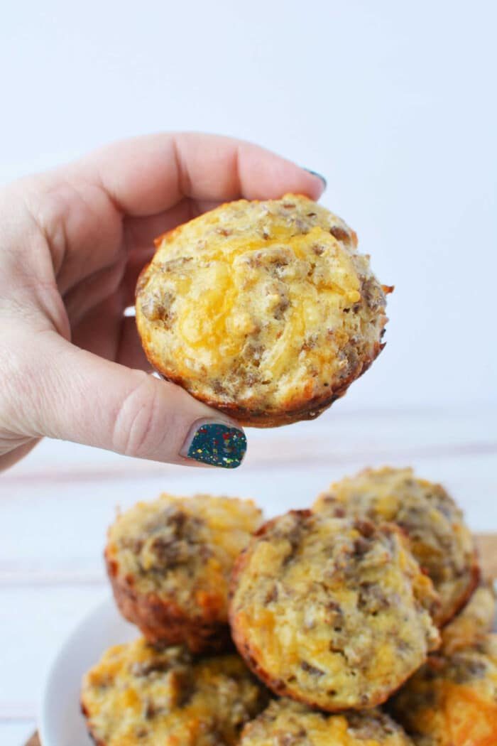 A hand holds a savory muffin crowned with melted cheese, its golden crust hinting at hidden delights within. In the background, a plate overflows with more enticing muffins, each potentially featuring hints of sausage for an added hearty twist. solid for those Bisquick-inspired morning feasts!.