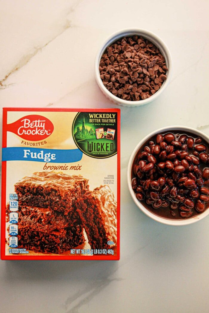 A box of fudge brownie mix sits alongside bowls of chocolate chips and black beans, perfectly set up to create good black bean brownies with mix on a pristine white surface.