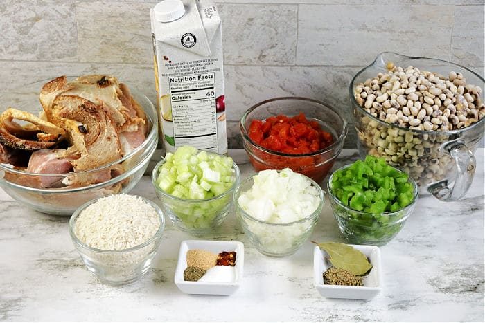 Ingredients for a soup or stew arranged on a counter, including black eyed peas and ham hocks, rice, vegetables, spices, broth, and meat.