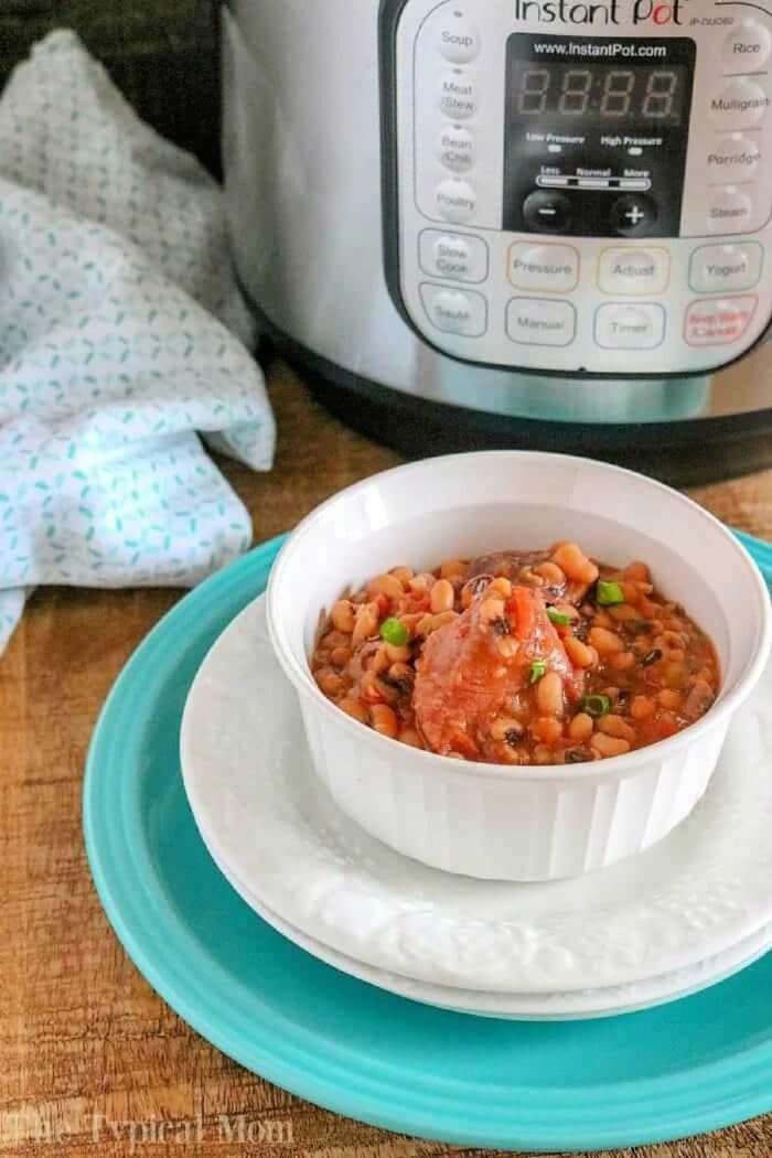 A white bowl of bean stew sits on plates in front of an Instant Pot, featuring hearty pressure cooker black eyed peas and ham, on a wooden surface.