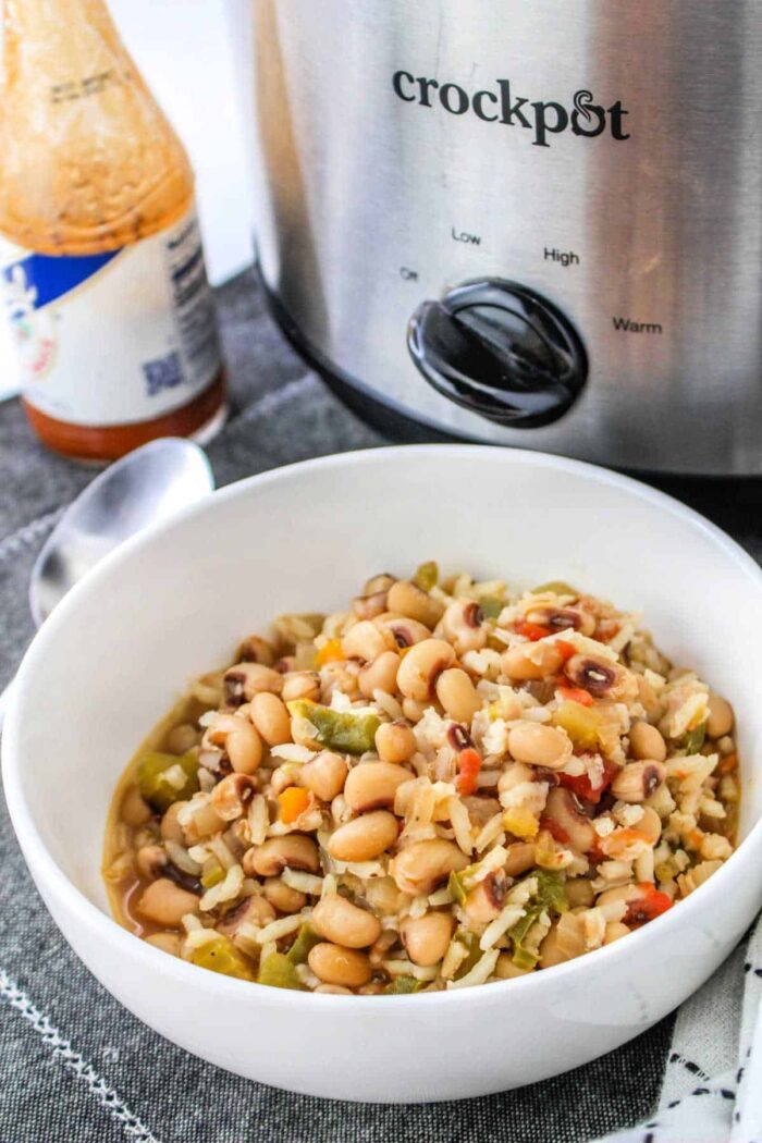 A bowl of Black Eyed Peas and Rice sits in front of a crockpot, accompanied by a spoon and a bottle of sauce, ready to enjoy.