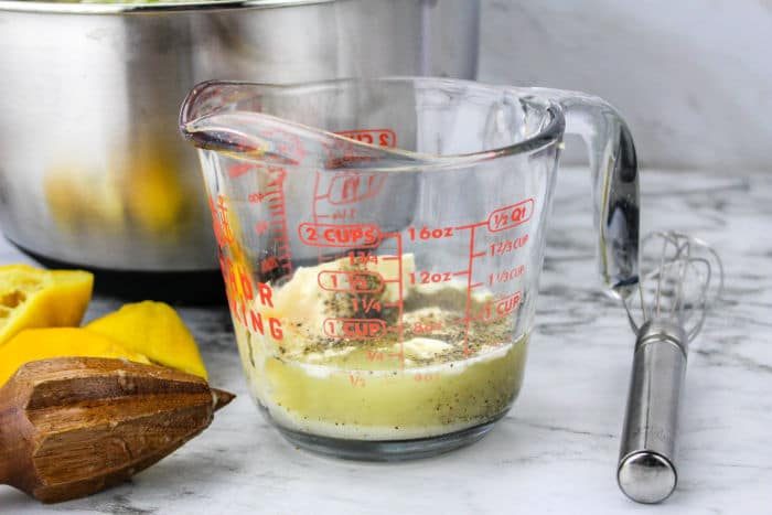 A glass measuring cup with coleslaw dressing sits on a marble surface, accompanied by a whisk, lemons, and a citrus reamer—solid for preparing tangy mixtures like Blue Cheese Coleslaw.