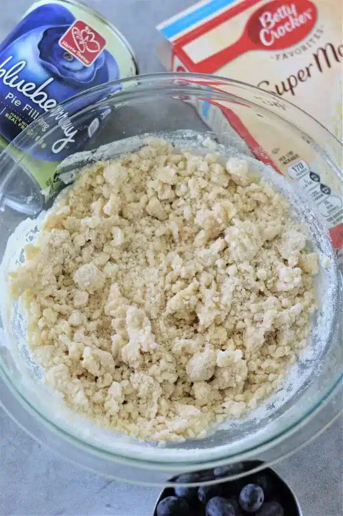 A glass bowl with crumbly dough mixture is surrounded by a blueberry pie filling can, a Betty Crocker Super Moist cake mix box, and a few scattered blueberries on a gray surface, hinting at the creation of a good blueberry cobbler with cake mix.