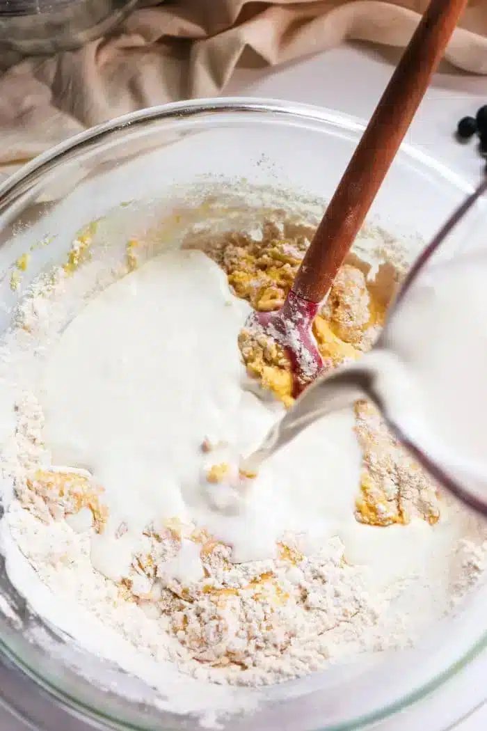 A glass mixing bowl contains flour and egg batter being mixed with a wooden spoon. A pitcher pours liquid, likely milk, into the bowl, solid for making blueberry muffin tops. The background shows part of a beige cloth and some blueberries on the table.