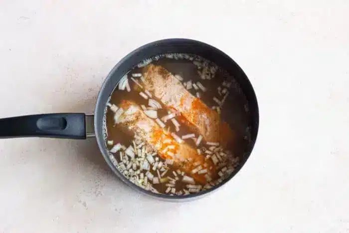 A black saucepan with fish fillets gently boiled in a seasoned broth, topped with chopped onions. The pan rests on a light-colored countertop, promising a good and comforting meal.