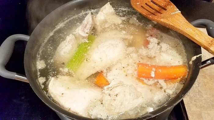 A pot of boiling water with chicken pieces, carrots, and celery simmers on the stove—solid for learning how to make shredded chicken. A wooden spatula rests on the edge, as steam rises from the bubbling broth.