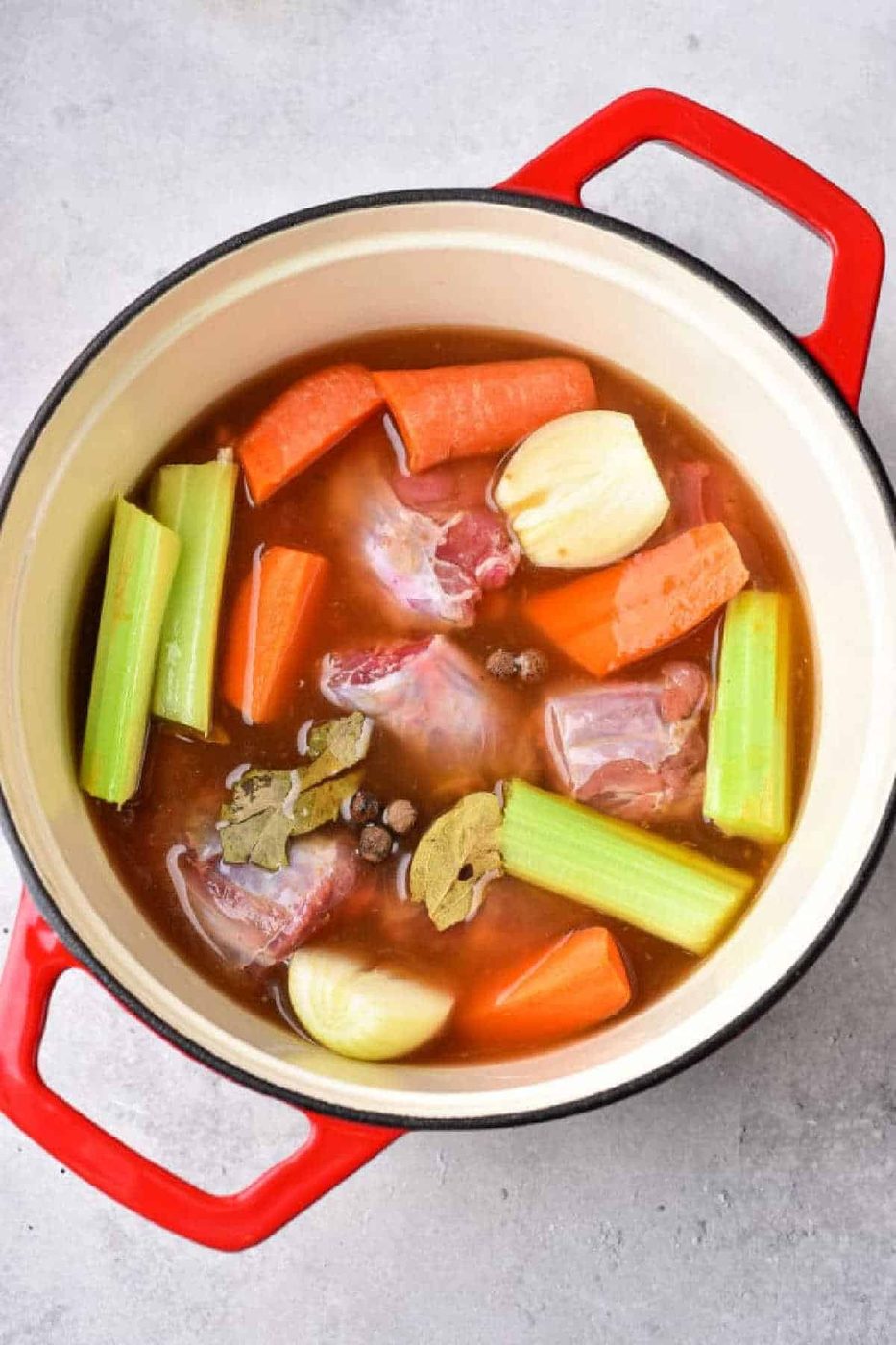 Red pot with raw beef, carrots, celery, onions, bay leaves, and broth—solid for making soups or pairing with boiled turkey necks—ready to cook on a gray surface.