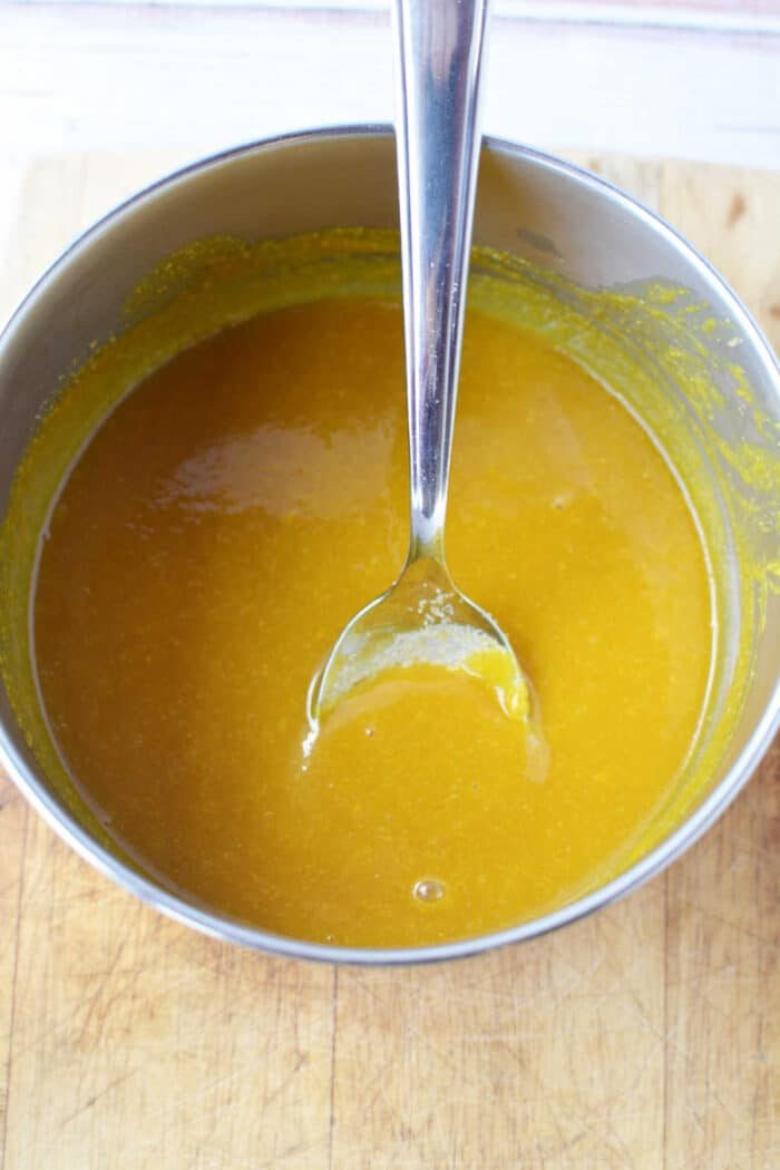 A metal bowl of yellow-orange Bologna Burnt Ends sauce with a spoon resting inside sits on a wooden surface.