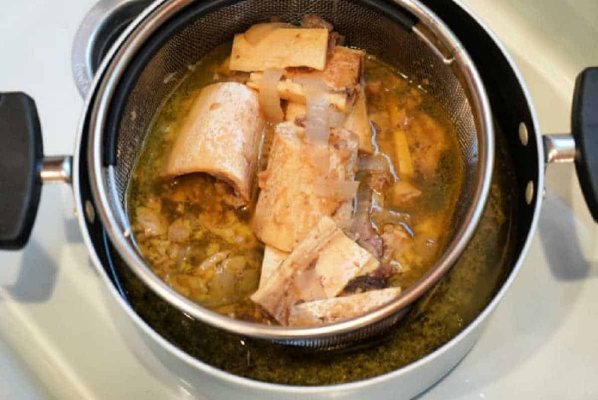 Large beef bones and vegetables in a strainer over a pot of broth on the stovetop—solid for preparing a nourishing Bone Broth Recipe Instant Pot enthusiasts will love.