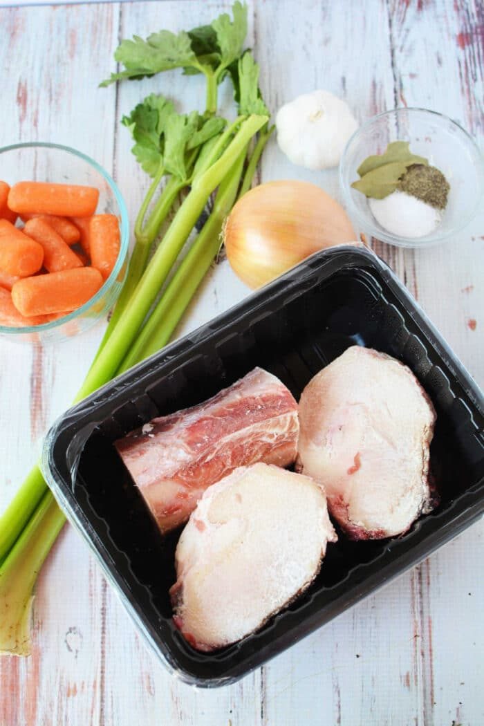 Ingredients for bone broth: celery, carrots, garlic, and onion lay next to beef bones on a tray. A bowl of spices with bay leaves completes the setup. Perfectly suited for a slow cooker, these elements promise depth and warmth in every sip.