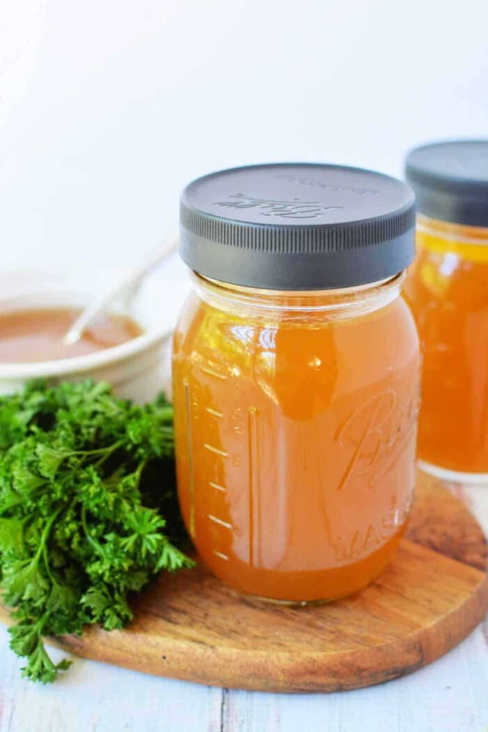 A mason jar of golden bone broth slow cooker on a wooden board, with fresh parsley and a bowl in the background.