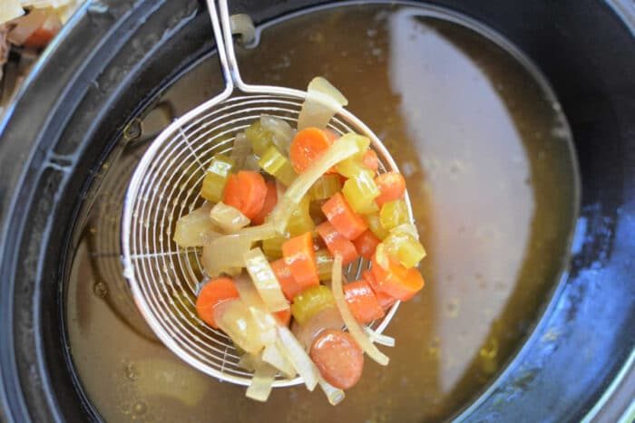 A wire ladle lifts diced carrots, celery, and onions above a pot of simmering bone broth, creating a fragrant medley solid for your slow cooker.