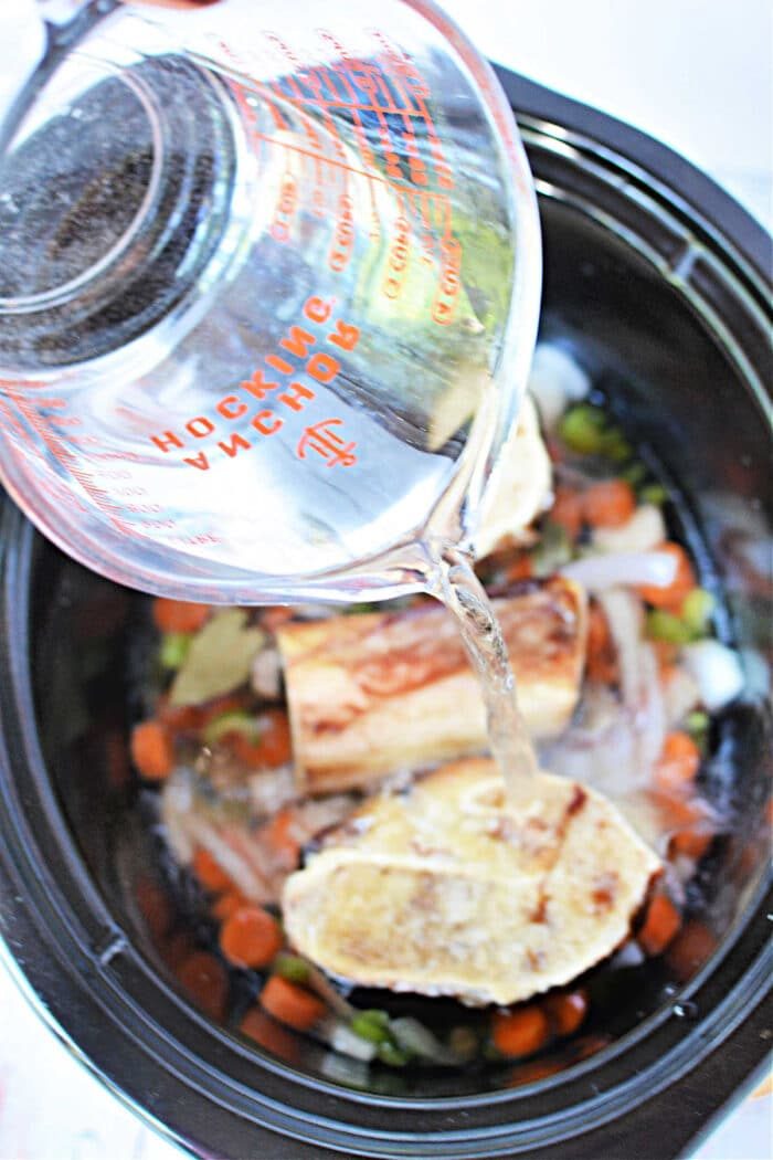 Pouring water into a slow cooker, creating a rich bone broth with vegetables and bones.