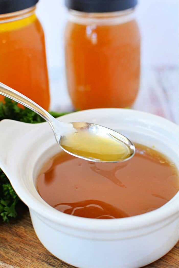 A spoon hovers over a white bowl, filled with homemade bone broth made in a slow cooker, with two jars in the background.