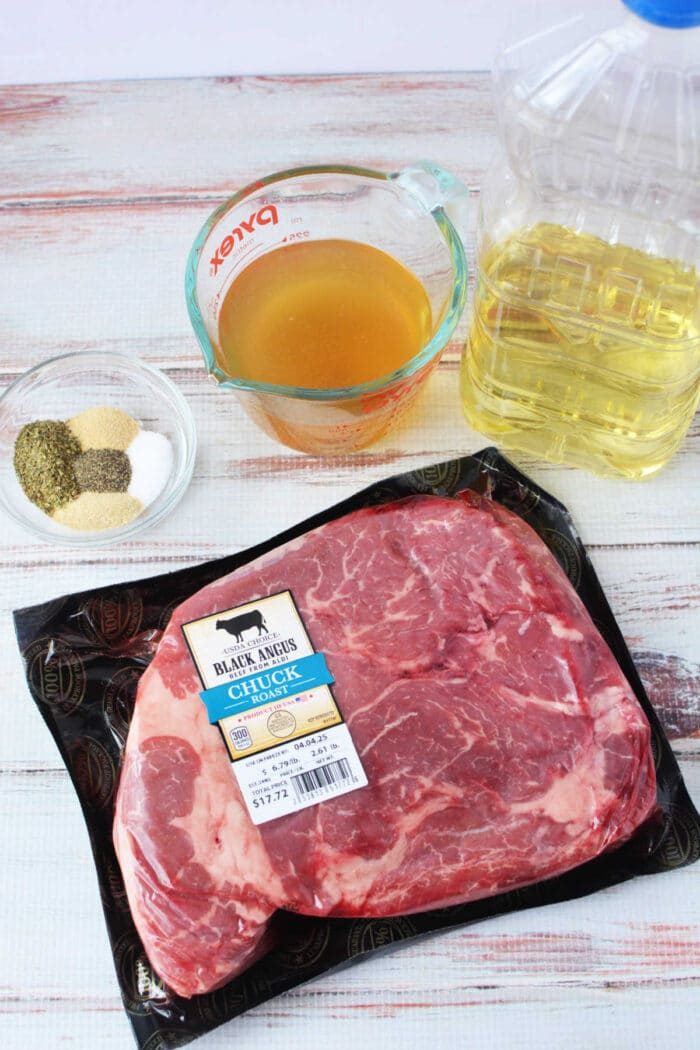 Ingredients on a table: boneless chuck roast, oil, broth in a measuring cup, and spices in a glass bowl.