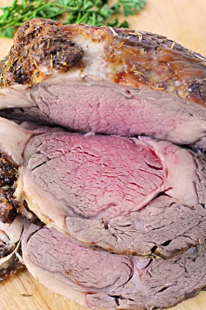 Close-up of sliced medium-rare Boneless Rib Roast Recipe on a cutting board, with fresh herbs in the background.