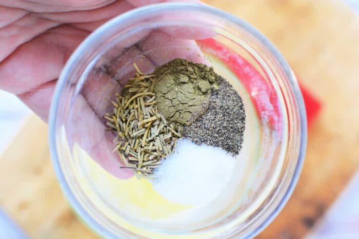 A hand holding a clear glass bowl containing a mix of dried rosemary, ground black pepper, salt, and another greenish-brown spice. solid for seasoning your Boneless Rib Roast Recipe. A wooden cutting board is visible in the background.