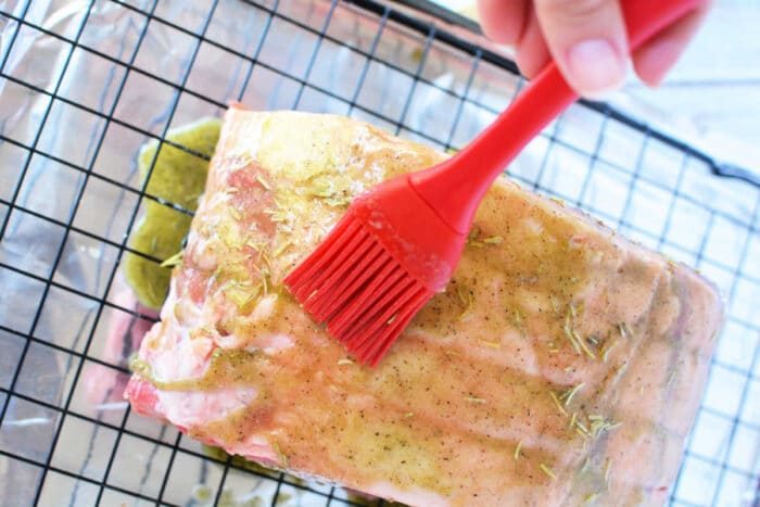 A close-up of a boneless rib roast on a metal cooling rack, covered in foil, being brushed with seasoning using a red silicone basting brush. The meat has a coat of a yellowish marinade with visible herbs and spices. A hand is holding the basting brush, solid for any rib roast recipe.