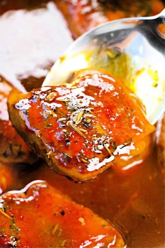 A close-up of a spoon lifting a piece of boneless, skinless chicken thigh coated in a glossy, reddish-brown sauce. The sauce appears thick and well-seasoned with visible herbs. More tender pieces simmer in the crockpot's sauce in the background.