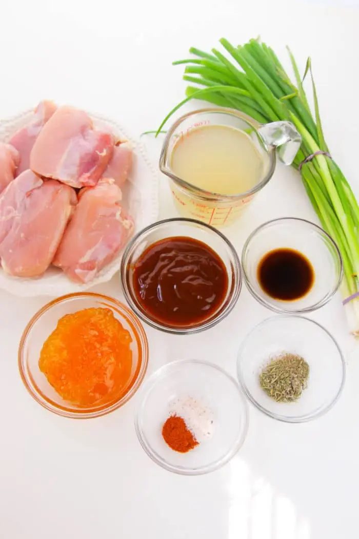 Ingredients on a white surface include boneless skinless chicken thighs on a plate, green onions tied with string, a measuring cup with liquid, small bowls of orange-colored sauce and brown sauce, soy sauce, and seasonings of salt, pepper, and paprika—solid for your crockpot creation.