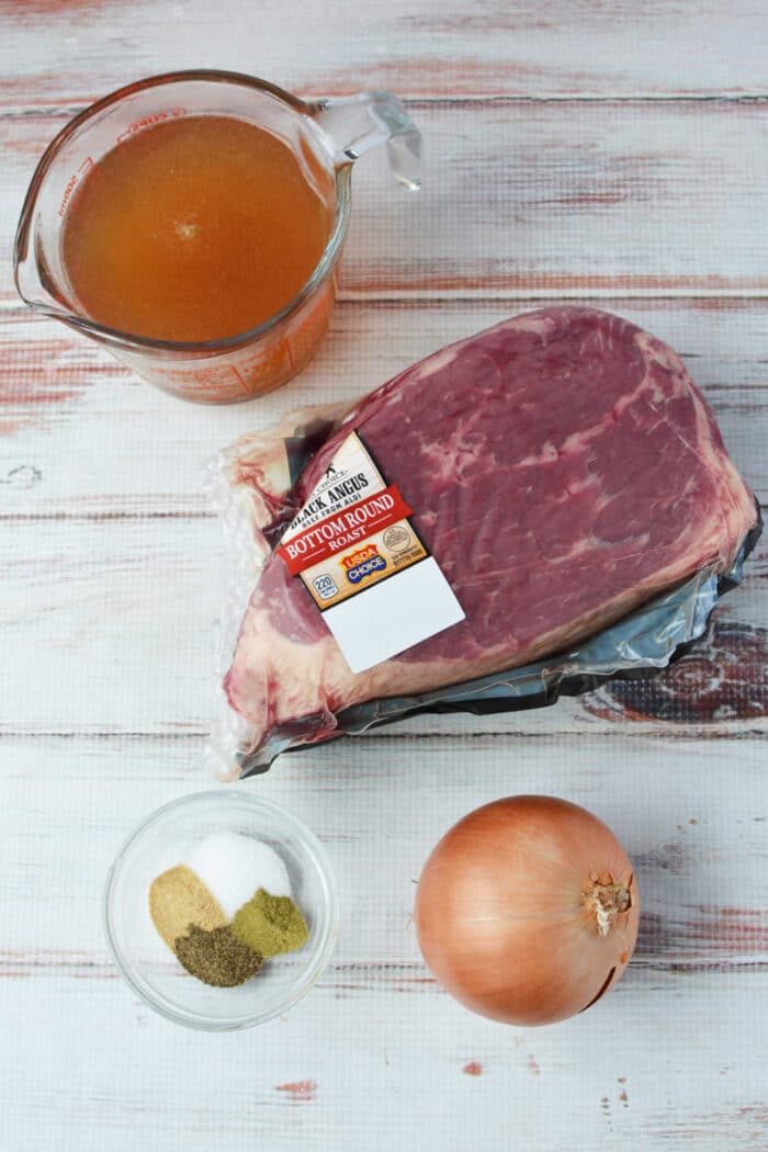 A raw beef bottom round roast, onion, spices, and beef broth in containers are arranged on a white wooden surface—solid for your next slow cooker meal.