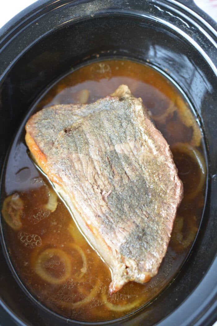 A piece of cooked Beef Bottom Round Roast sits in broth with onions inside a black slow cooker.