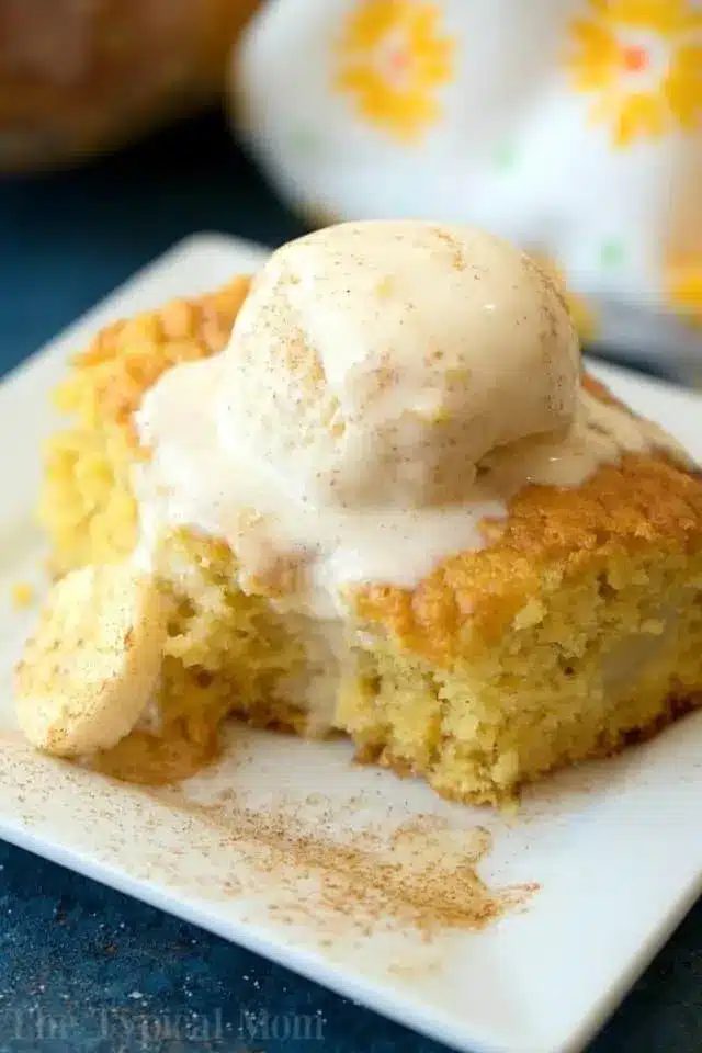 A slice of cake, made using clever box cake mix hacks, is topped with a scoop of vanilla ice cream on a white square plate. The cake boasts a light golden color, with a small cookie on the side. Cinnamon graces the top and surrounds the dessert, set against a backdrop of patterned cloth.