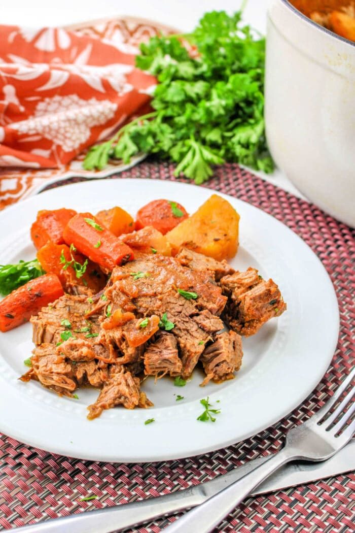 Plate of savory pot roast with carrots and potatoes, braised to perfection, garnished with herbs on a woven mat, and parsley in the background.