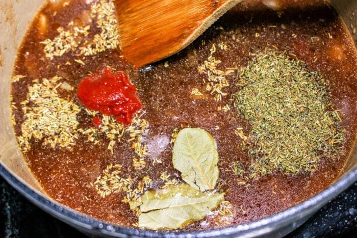 A cooking pot simmers with rich broth, herbs, bay leaves, and tomato paste. A wooden spoon stirs the ingredients into a fragrant harmony, ideal for braising with chuck roast and a splash of red wine.